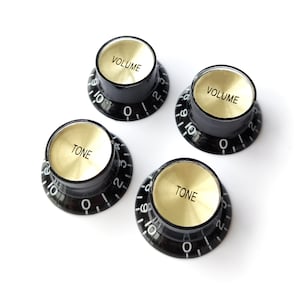 Pode incluir: Quatro botões de guitarra pretos com topos dourados. Os botões são etiquetados como "VOLUME" e "TONE".