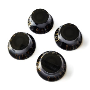 Pode incluir: Quatro botões de guitarra pretos com números e marcas douradas. Os botões são redondos com um centro elevado e um fundo plano.