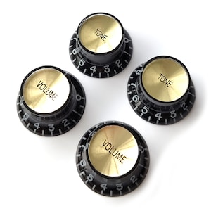 Pode incluir: Quatro botões de controle de guitarra pretos com tampas douradas. Dois botões estão rotulados como "VOLUME" e dois como "TONE". Cada botão tem uma escala numerada de 0 a 10, com números brancos sobre fundo preto.