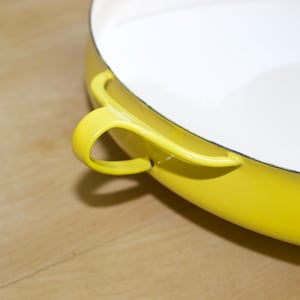 Puede incluir: Una olla de hierro fundido esmaltada amarilla con interior blanco y una sola asa.