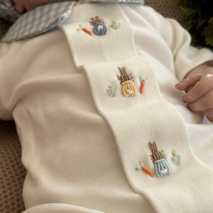 May include: A white baby onesie with a light blue gingham collar and embroidered bunny and carrot designs. The onesie features a decorative bib with embroidered details. The overall design is soft and pastel-coloured.