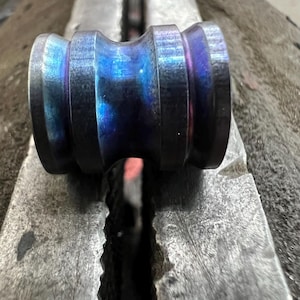 May include: A cylindrical metal bead with a concave centre and a heat-treated finish displaying iridescent blue and purple hues. The object features a smooth, polished surface with distinct circular ridges at both ends, creating a spool-like shape.