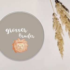 Op de afbeelding: Een rond, grijs, decoratief bord met de tekst "grosser Bruder" in witte, cursieve letters. Een cartoon-leeuw met oranje vacht en een roze neus is gecentreerd onder de tekst.