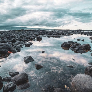 Può includere: Una pozza d'acqua calma e riflettente circondata da rocce vulcaniche scure. Il cielo è coperto di nuvole che si riflettono nell'acqua.