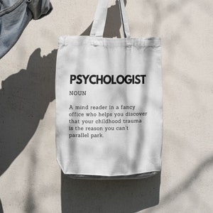 May include: White tote bag with the word "PSYCHOLOGIST" printed in black. Below the word is a definition. The bag is held by a person's hand against a neutral background.