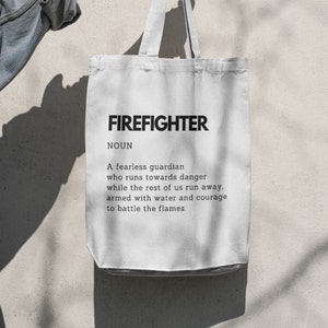 Könnte beinhalten: Weißer Canvas-Beutel mit dem Wort "FIREFIGHTER" in fetter schwarzer Schrift und einer Definition. Der Beutel wird von einer Person gehalten. Die Definition beschreibt einen furchtlosen Wächter.
