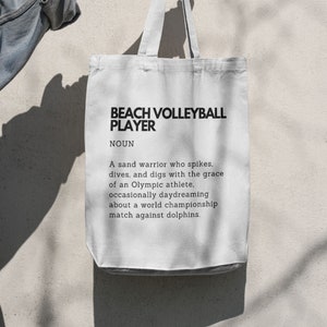 May include: White canvas tote bag with black text that reads "BEACH VOLLEYBALL PLAYER NOUN A sand warrior who spikes, dives, and digs with the grace of an Olympic athlete. occasionally daydreaming about a world championship match against dolphins."