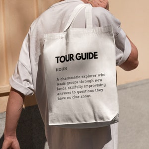 Peut inclure: Un sac fourre-tout en toile blanche avec du texte noir qui dit "TOUR GUIDE NOUN A charismatic explorer who leads groups through new lands, skillfully improvising answers to questions they have no clue about."