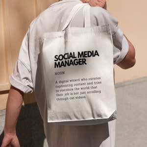 May include: White tote bag with black text that reads "SOCIAL MEDIA MANAGER NOUN A digital wizard who curates captivating content and tries to convince the world that their job is not just scrolling through cat videos."