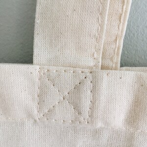 May include: Close-up of a white canvas tote bag with a reinforced stitched seam at the top of the bag.