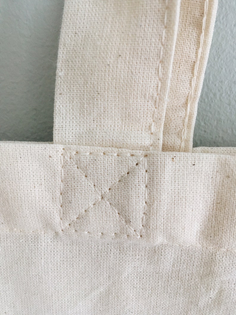 May include: Close-up of a white canvas tote bag with a reinforced stitched seam at the top of the bag.