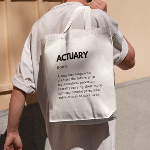 May include: White tote bag with the word "ACTUARY" in bold black letters. Below, the definition of actuary is printed in black. The bag is carried over the shoulder of a person wearing a light gray t-shirt.