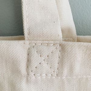 May include: Close-up of the top of a white canvas tote bag with a reinforced seam.