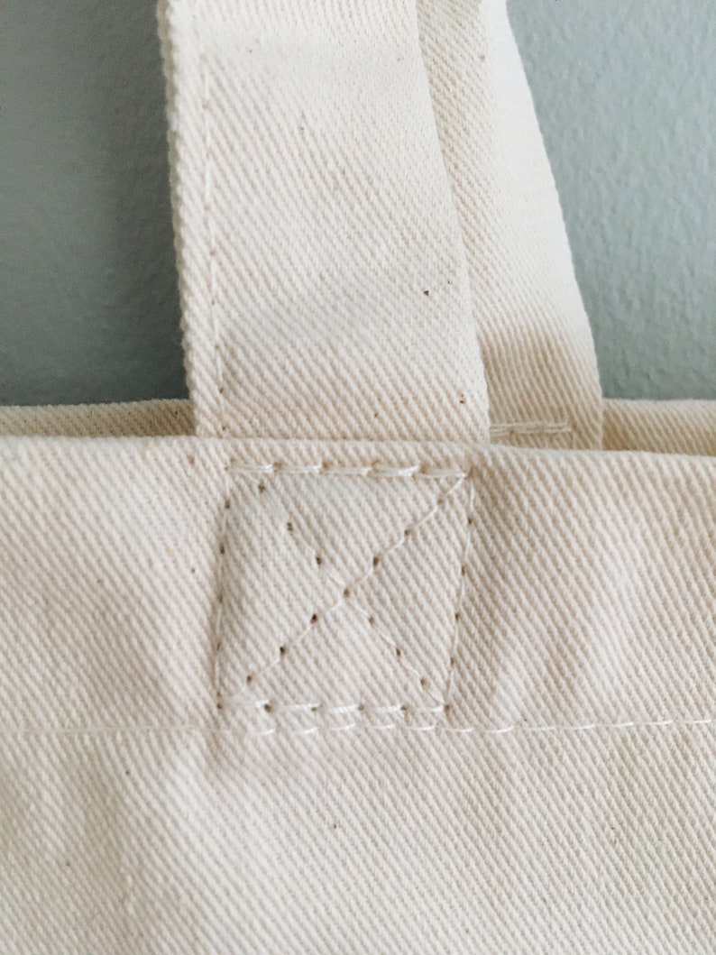 May include: Close-up of the top of a white canvas tote bag with a reinforced seam.