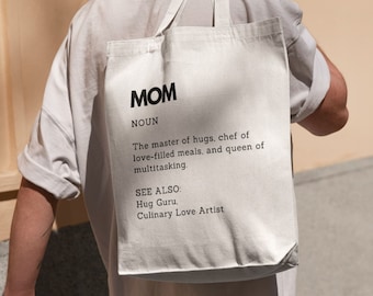 May include: White tote bag with black text that reads "MOM NOUN The master of hugs, chef of love-filled meals, and queen of multitasking. SEE ALSO: Hug Guru. Culinary Love Artist"