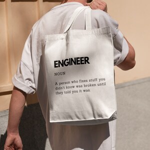 May include: Cream-coloured tote bag featuring the word "ENGINEER" in black, with a definition below. The bag is carried over a person's shoulder. The definition is printed in black.