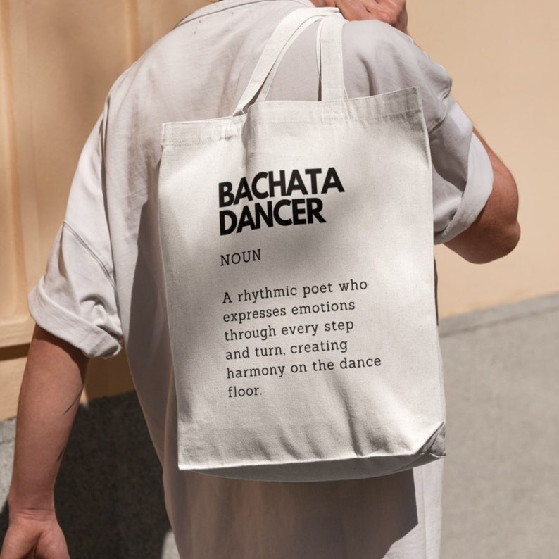 May include: A white tote bag with black text that reads "BACHATA DANCER" and a definition of the term. The definition reads "A rhythmic poet who expresses emotions through every step and turn, creating harmony on the dance floor."