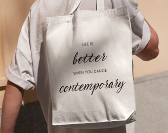 May include: A beige tote bag with black text that reads "LIFE IS better WHEN YOU DANCE contemporary." The bag has two handles and is made of a sturdy fabric. The bag is being carried over a person's shoulder.