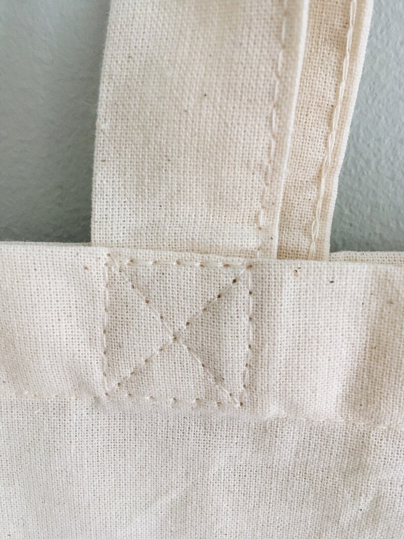 May include: Close-up of a natural canvas tote bag. The bag features a sturdy, cream-colored fabric with visible texture. The handle is securely stitched to the bag with reinforced stitching in an X pattern, ensuring durability. The overall design is simple and functional.