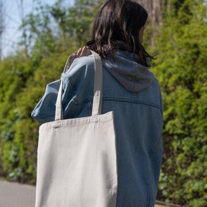 Peut inclure: Une personne portant une veste en jean et un sweat-shirt gris porte un grand sac fourre-tout en toile blanche.