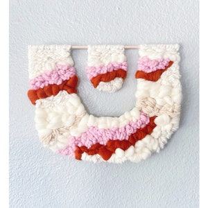 May include: A wall hanging made of woven yarn in white, pink, and orange. The hanging is in the shape of a U with two smaller U-shaped pieces hanging above it.