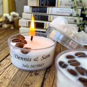 May include: A burning candle in a clear plastic container, adorned with coffee beans. The candle is inscribed with "Dennis & Sam July 26th, 2025". The candle sits on a wooden surface.