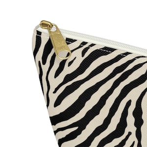 May include: A cosmetic bag with a zebra print pattern in black and cream. The bag has a gold-colored zipper and a white zipper pull. The bag is made of a durable fabric.