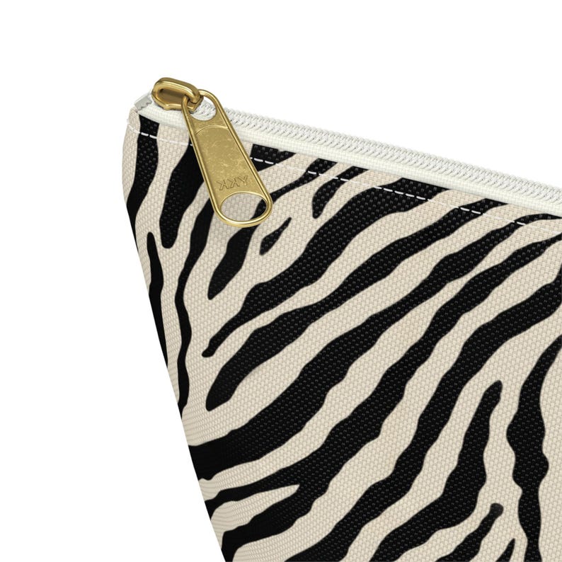 May include: A cosmetic bag with a zebra print pattern in black and cream. The bag has a gold-colored zipper and a white zipper pull. The bag is made of a durable fabric.