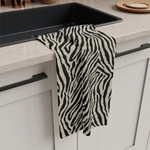 May include: A black and beige zebra print kitchen towel hangs over a countertop. The towel is made of a textured fabric and features a bold, repeating pattern. The towel is near a black sink and white cabinets.