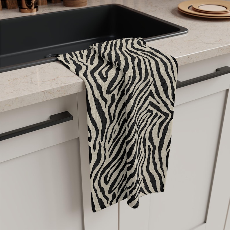 May include: A black and beige zebra print kitchen towel hangs over a countertop. The towel is made of a textured fabric and features a bold, repeating pattern. The towel is near a black sink and white cabinets.