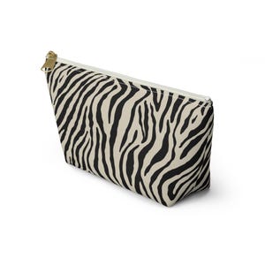 May include: A beige and black zebra print cosmetic bag with a gold zipper. The bag is rectangular with a trapezoid shape. The zipper runs along the top edge. The bag is made of a soft, durable material.