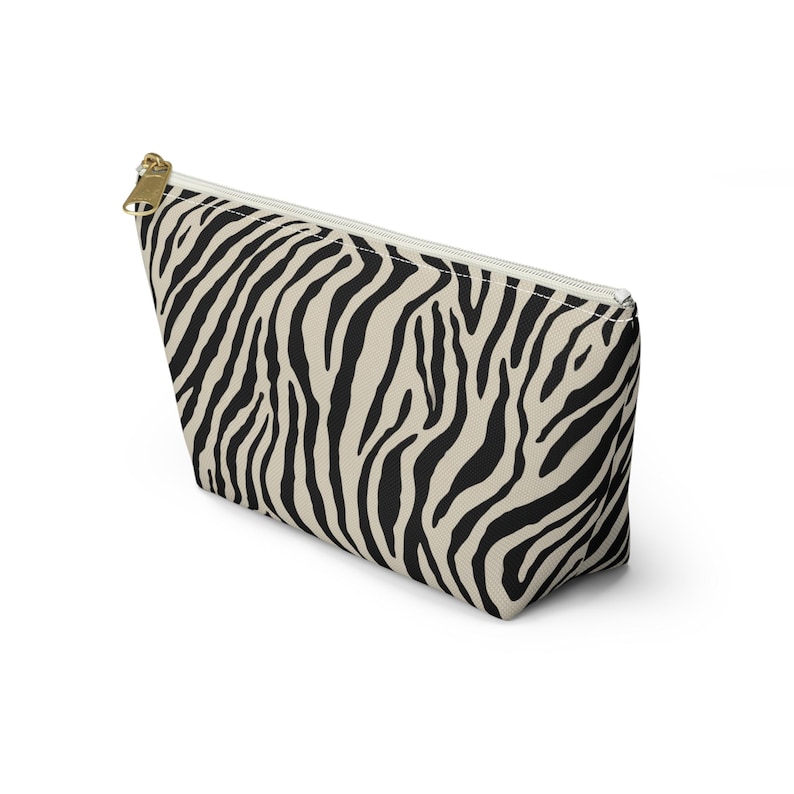 May include: A beige and black zebra print cosmetic bag with a gold zipper. The bag is rectangular with a trapezoid shape. The zipper runs along the top edge. The bag is made of a soft, durable material.