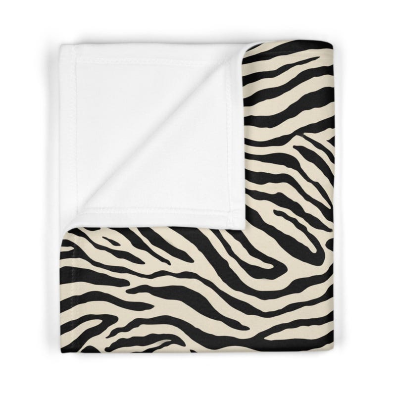 May include: A folded blanket with a zebra print design. The blanket features a cream-colored background with black stripes. The edge of the blanket is white.