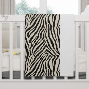 May include: A baby blanket with a black and beige zebra print. The blanket is draped over a white crib. The crib is in front of a window with white curtains. The blanket is made of soft material.