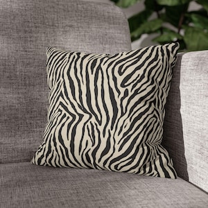 May include: Square decorative pillow with a black and cream zebra stripe pattern. The pillow is placed on a gray upholstered chair, offering a bold accent for home decor. The pillow's design adds a touch of wild style to any living space.