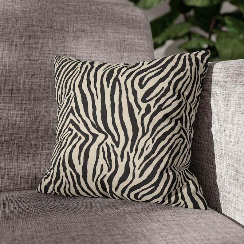 May include: Square decorative pillow with a black and cream zebra stripe pattern. The pillow is placed on a gray upholstered chair, offering a bold accent for home decor. The pillow's design adds a touch of wild style to any living space.