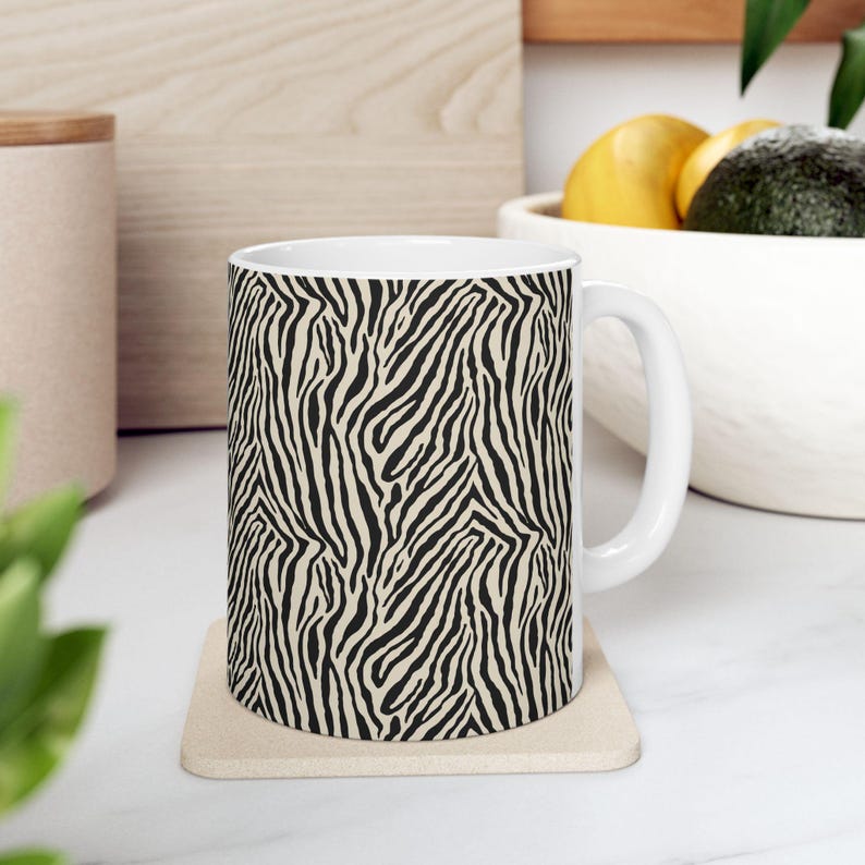 May include: White ceramic coffee mug with a black and beige zebra stripe pattern. The mug has a white handle and sits on a beige coaster. The mug is filled with a dark liquid.