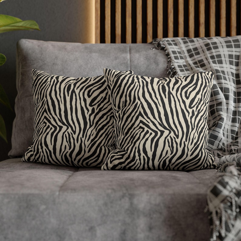 May include: Two square throw pillows with a black and cream zebra stripe pattern. The pillows are placed on a gray sofa, with a plaid blanket visible in the background. The pillows add a touch of animal print to home decor.