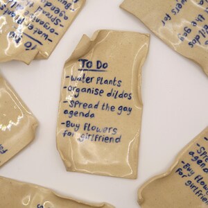 May include: A collection of beige ceramic tiles with blue handwritten text. The central tile reads "To Do" with a list: "Water Plants, organise dildos, Spread the gay agenda, Buy Flowers for girlfriend."
