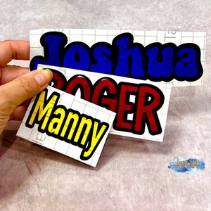 May include: Three vinyl stickers with names "Joshua", "Roger", and "Manny" in bold, colorful letters. The stickers are on a white gridded background.