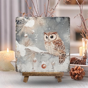 May include: A square decorative plaque featuring a brown and white owl perched on a snowy pine branch. The background is a soft gray watercolor wash with white snowflakes, pine needles, and small hanging ornaments in shades of orange and gold.
