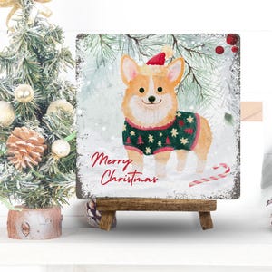 May include: A square decorative sign featuring a watercolor illustration of a tan and white Corgi wearing a red Santa hat and a green sweater with red tree patterns. The text Merry Christmas is written in red script. A candy cane lies on the snow.