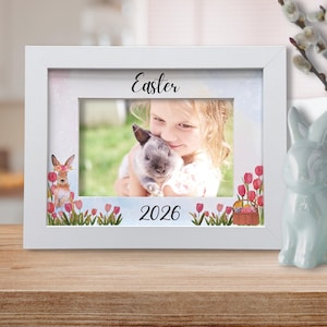 May include: White framed photo with the word "Easter" and the year "2026". The photo features a child holding a bunny. Decorative bunny and eggs are on the table.
