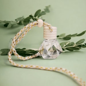 May include: A clear glass bottle with a white wooden lid and a gold and white string attached. The bottle is shaped like a diamond and is filled with a clear liquid. The bottle is hanging from a green leafy branch.