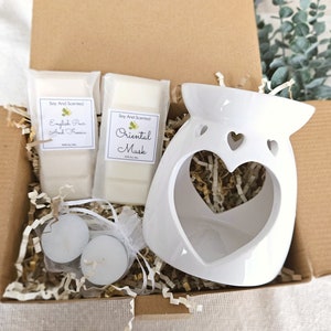 May include: A white ceramic heart-shaped wax warmer with two soy wax melts, one labelled "English Pear and Freesia" and the other "Oriental Musk", and two tea lights in a white organza bag.