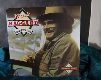 Pode incluir: Uma capa de álbum de vinil vintage com Merle Haggard, um cantor e compositor de música country. A capa apresenta uma foto de perto de Haggard com um chapéu marrom e uma jaqueta verde, segurando um violão. O título do álbum é "Merle Haggard Songwriter" e faz parte da "Songwriter Series".