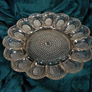 May include: A clear glass egg serving platter. It has a central circular area and twelve individual egg-shaped compartments around the edge. The platter has a textured, dotted pattern in the centre and in each egg cup. The edges are scalloped.