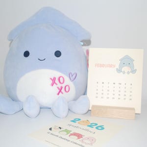 May include: A light blue plush squid toy with embroidered "XO XO" and a heart. A calendar for February 2026 and a 12-month desktop calendar are also present. The calendar features a squid illustration.