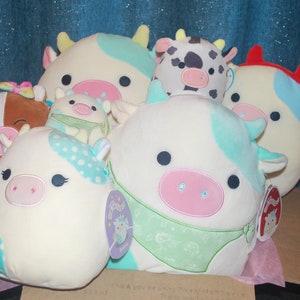 May include: A collection of plush cow toys in various colors and patterns. The cows have soft, rounded bodies, with details like pink noses, black eyes, and pastel-colored accents. Some have polka dots, and others have bandanas. The toys are arranged in a cardboard box.