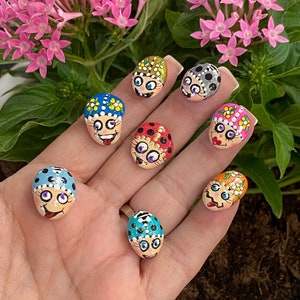 May include: A collection of hand-painted, whimsical face-shaped stones with various colorful designs. Each stone features a unique expression and decorative elements like polka dots and floral patterns. The stones are displayed against a backdrop of pink flowers and green foliage.